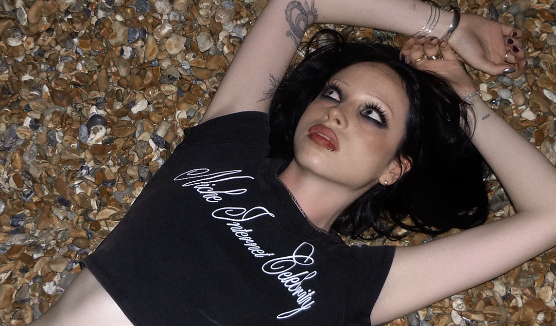 A woman lying on a bed of small brown pebbles, wearing a black cropped t-shirt with white script text that reads 'Niche Internet Celebrity'.