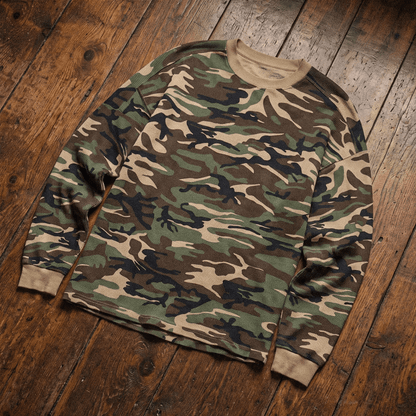 A long-sleeve crewneck sweatshirt in a classic woodland camouflage pattern, laid flat on a rustic dark wood plank floor.
