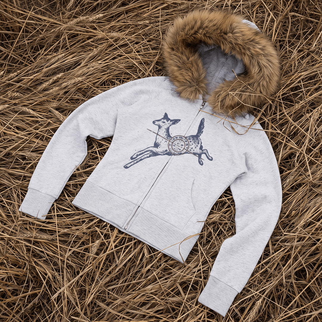 A light gray zip-up hoodie with a brown faux-fur trimmed hood, laid flat on a bed of dry straw. The front features a graphic of a jumping deer with a red crosshair pattern.
