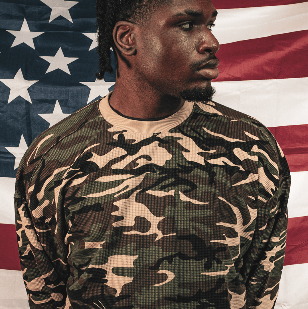 A man wearing a long-sleeve crewneck sweatshirt with a green, black, and beige camouflage pattern, posed in front of an American flag.
