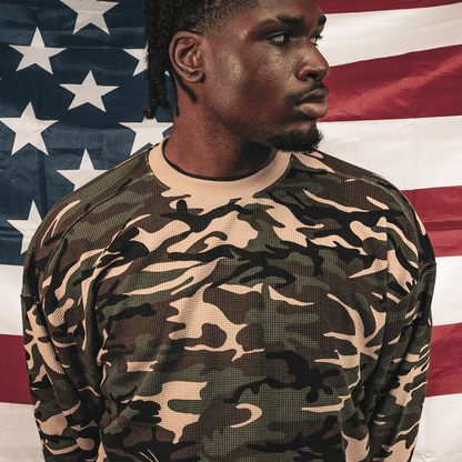 A man wearing a long-sleeve crewneck sweatshirt with a green, black, and beige camouflage pattern, posed in front of an American flag.