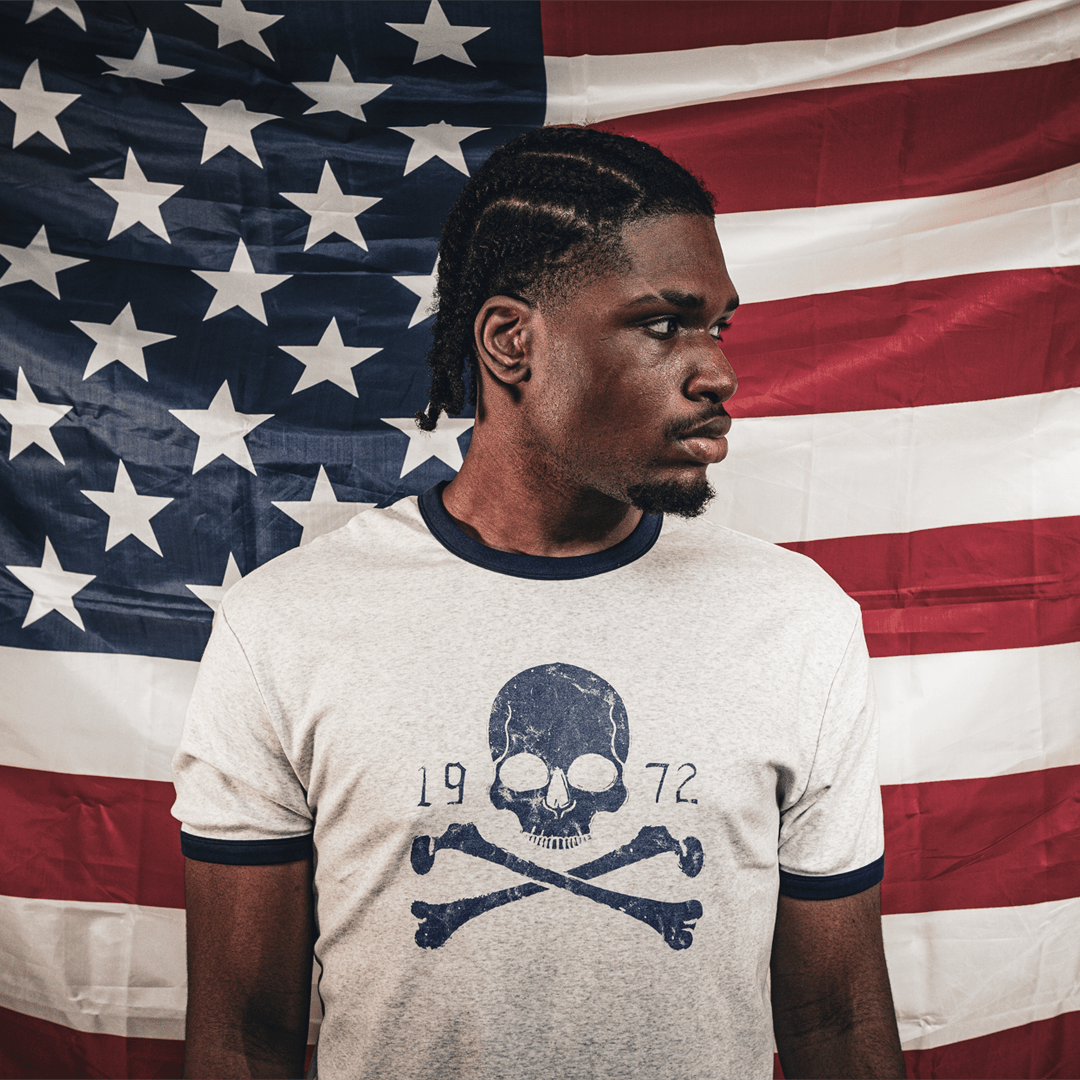 A man wearing a light gray heather t-shirt with a navy blue skull and crossbones graphic, posed in front of a large American flag backdrop.